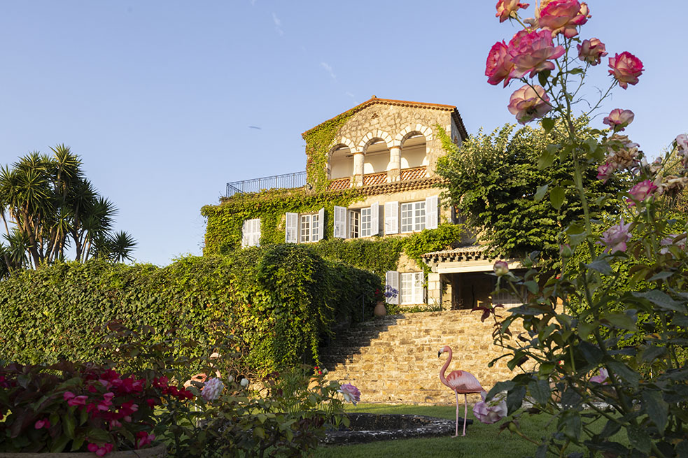 La facciata del mas provenzale in pietra che guarda verso la baia di Golfe-Juan. Una grande scalinata conduce alla terrazza sottostante: al centro del prato, una piccola fontana in pietra decorata con due fenicotteri. In primo piano, arbusti di rose.